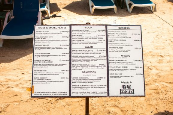 Menu board displayed on the beach, listing food and drink options.